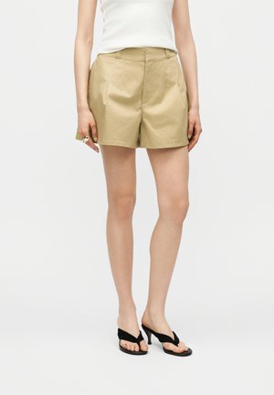 Kvinde iført beige højtaljede plisserede shorts, hvid ærmeløs top, sorte hælede sandaler og en guldring på venstre hånd.