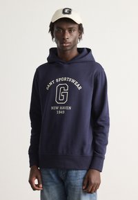 Felpa blu marina con grafica frontale che presenta "GANT SPORTSWEAR", "NEW HAVEN" e "1949" in carattere crema; abbinata a un cappellino beige chiaro.