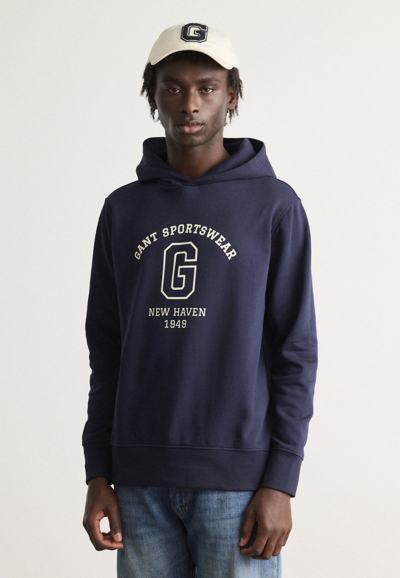 Felpa blu marina con grafica frontale che presenta "GANT SPORTSWEAR", "NEW HAVEN" e "1949" in carattere crema; abbinata a un cappellino beige chiaro.