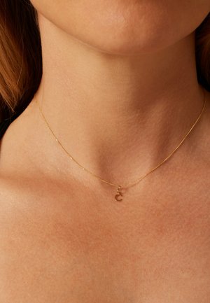 Gold chain necklace with a fine, delicate texture, featuring a small, round initial pendant. Worn close to the neck.