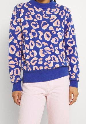 Pull bleu avec des taches léopard roses et blanches, porté avec un pantalon rose clair, visible du milieu de la poitrine jusqu'aux hanches.