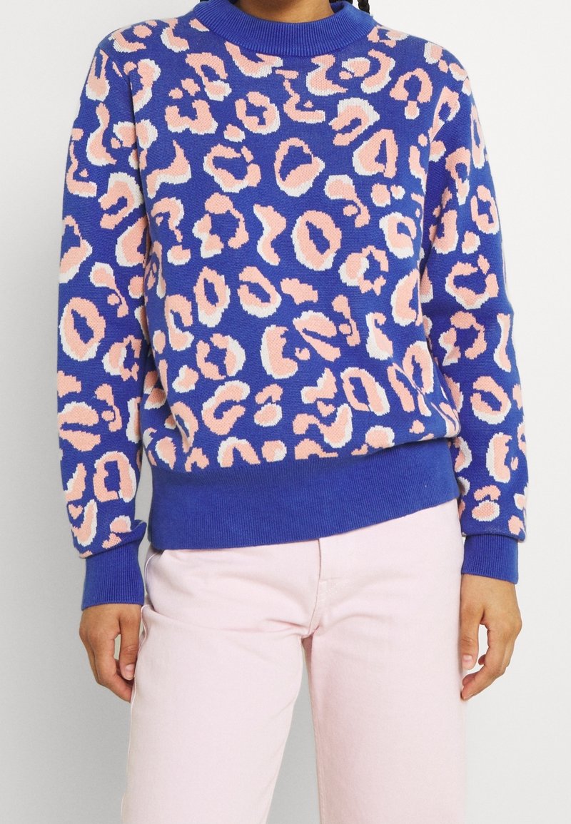 Pull bleu avec des taches léopard roses et blanches, porté avec un pantalon rose clair, visible du milieu de la poitrine jusqu'aux hanches.
