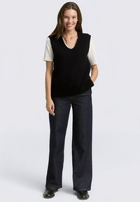 Gilet en maille côtelée noire avec col en V, porté sur un t-shirt blanc. Associé à un jean large foncé et des chaussures noires. Design simple, coupe décontractée.