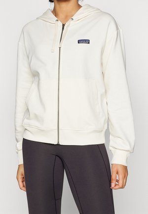 Donna che indossa una felpa bianca con zip Patagonia con tasche frontali e leggings neri, in piedi davanti a uno sfondo semplice.