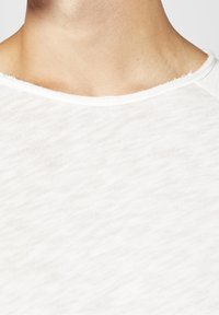 Vit, lättviktsmaterial med en något texturerad yta. Rund halsringning med fransade kanter, vilket visar subtiliteter i vävstruktur och färg.