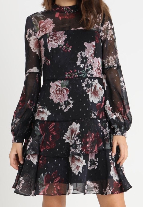Robe noire transparente à manches longues avec imprimé floral rose et bordeaux, jupe à volants superposés et col montant, portée à mi-cuisse.