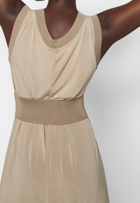 Robe beige sans manches avec des accents en tricot côtelé au niveau du col et de la taille, présentant un tissu lisse et fluide ainsi qu'un détail de jupe plissée.