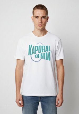 Jeune homme portant un t-shirt blanc Kaporal Denim et un jean bleu, debout devant un fond clair uni.