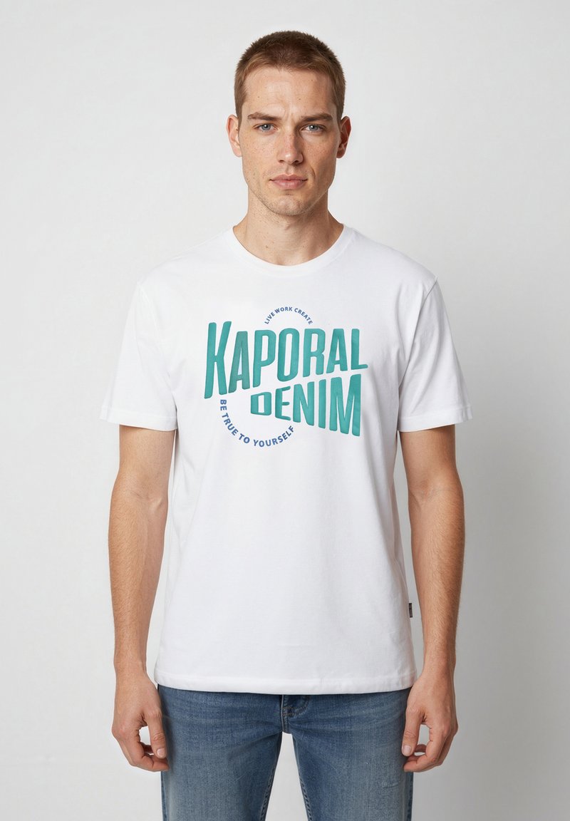 Jeune homme portant un t-shirt blanc Kaporal Denim et un jean bleu, debout devant un fond clair uni.