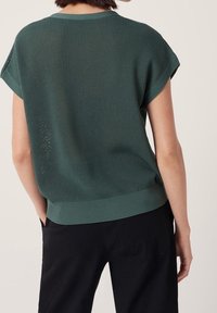 Donna che indossa un maglione corto a maniche corte di un verde scuro testurizzato e pantaloni neri, mostrata di spalle con una mano in tasca.