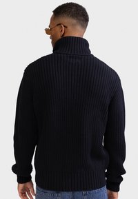 Gestrickter schwarzer Rollkragenpullover mit Rippstruktur und Bündchen, mit lockerer Passform und hohem Kragen. Rückansicht hebt das Design des Pullovers hervor.