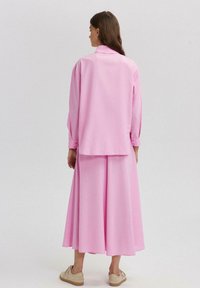 Růžový dvoudílný outfit obsahující dlouhý rukáv a splývavou sukni. Materiál je hladký, s jemnou strukturou, a doplněný béžovými teniskami.