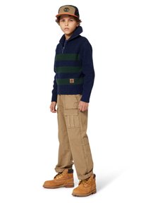 Navy und grün gestreifter Strickpullover mit hohem Kragen, hellbraune Cargo-Hosen und braune Arbeitsschuhe. Die Mütze hat ein Logo mit einem Baumdesign.