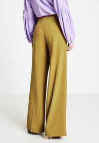 Weit geschnittene Hosen aus olivgrünem Stoff mit hoher Taille und klaren Linien. Kombiniert mit einer lavendelfarbenen Bluse mit gepufften Ärmeln.