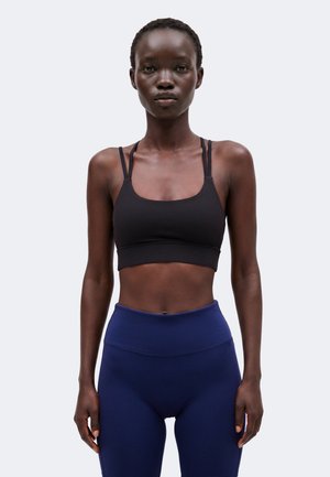 Donna atletica che indossa un reggiseno sportivo nero e leggings blu navy a vita alta, in piedi davanti a uno sfondo chiaro e uniforme.