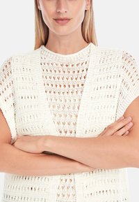 Cremfarbener gehäkelter Cardigan mit kurzen Ärmeln, der offen gearbeitete Muster und eine lockere, entspannte Passform bietet. Weiche Textur und leichtes Design.