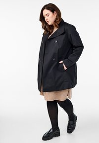 Cappotto nero con ampio risvolto, design doppiopetto e due tasche laterali. Indossato sopra un vestito beige e abbinato a mocassini neri.