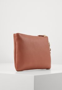 Pochette en cuir marron texturé avec fermeture éclair, présentant une finition lisse et un petit accent en matière dorée sur le côté.