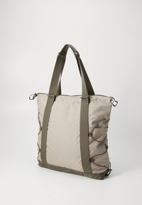 Beige stoffen draagtas met groene handvatten, verstelbare zijknoopjes en een ritssluiting aan de bovenkant. Glad textuur en een ruime ontwerping voor gemakkelijk vervoer.