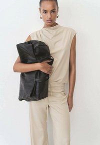Black leather duffel bag with textured surface, held in arms; minimalistic beige outfit with short sleeves and drawstring waist. White background.