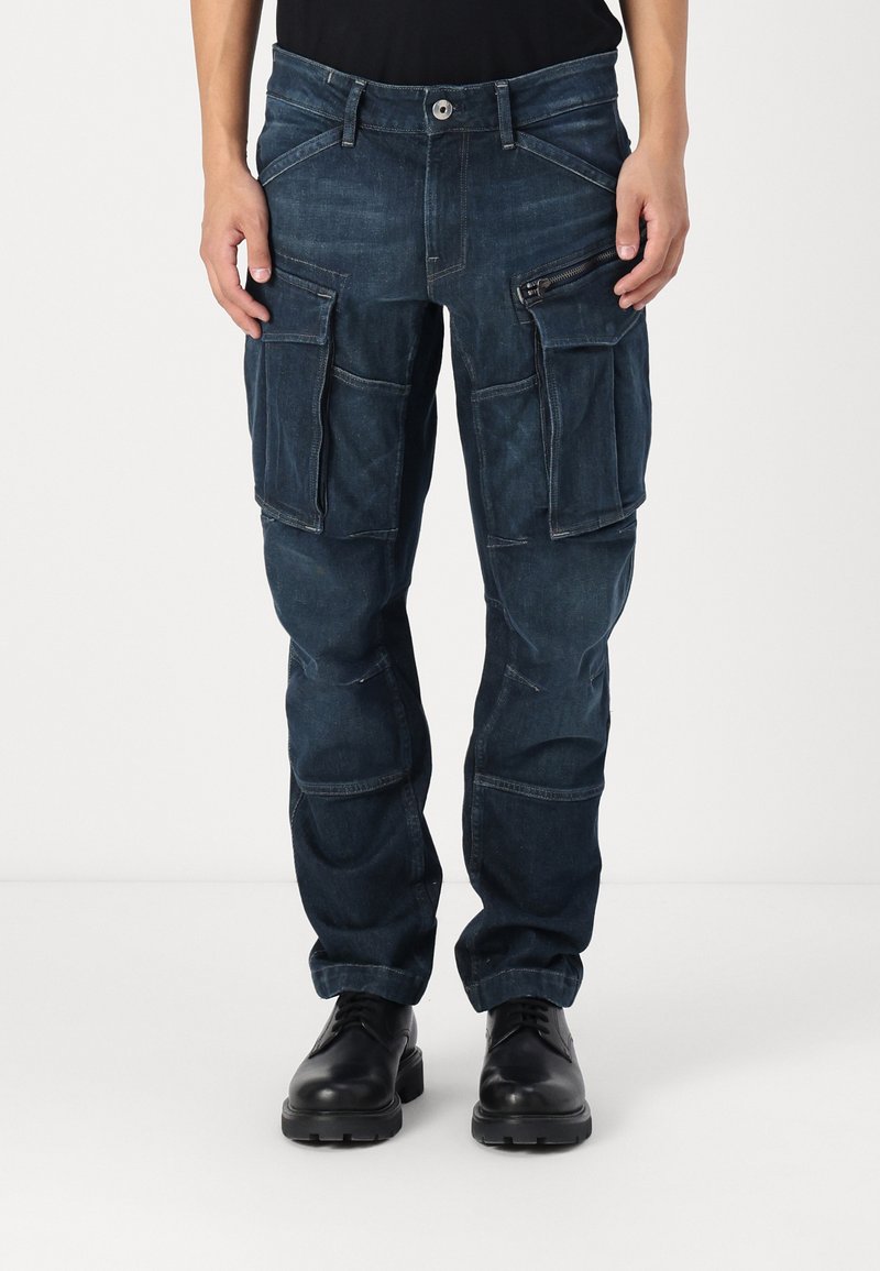 Dark blue cargo pants made of denim, featuring multiple large pockets, a zippered pocket, and a tapered leg design.