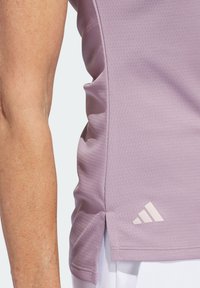 Jasnofioletowy top sportowy z fakturowanej tkaniny, z bocznym rozcięciem i małym białym logo Adidas na dolnym brzegu.