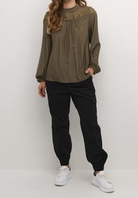 Blouse olive verte à manches longues avec des détails en dentelle aux épaules et un devant plissé, associée à un jogging noir à poignets élastiques et des baskets blanches.
