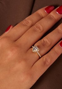 Gouden ring met een hartvormige, heldere edelsteen in het midden, omgeven door kleinere stenen op een gestructureerde band. Gepolijste rode nagels zichtbaar.