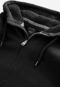 Sort zip-up hoodie med fleeceforet indvendigt. Har en glat overflade, snore til justering og en sølvfarvet lynlås. Mærket "NEXT."