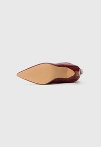 Zapato de tacón alto de cuero burdeos con punta afilada, mostrado desde el lado de la suela sobre un fondo blanco.