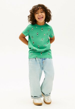 Smilende barn med krøllet hår iført en grøn t-shirt med basketballspiller-tryk, lyseblå jeans og hvide sneakers mod en hvid baggrund.