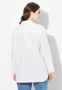 Camicia bianca a maniche lunghe con una piega sul retro, polsini con bottoni e un sottile ricamo floreale sul collo. Abbinata a jeans blu.