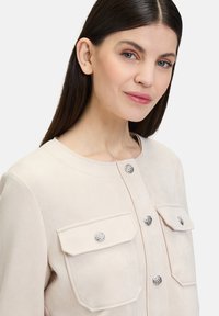 Beige Wildlederjacke mit rundem Ausschnitt, silbernen Druckknöpfen und zwei vorderen Taschen mit dekorativem Zubehör. Glatte Textur.