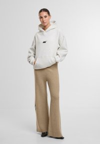 Svetle šedá mikina s černým logem, přední kapsou a šňůrkou na stažení. V kombinaci s volnými, texturovanými béžovými kalhotami a černými botami.