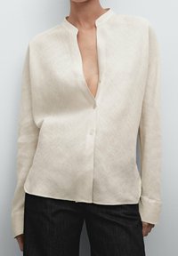 Kræmfarvet linnedskjorte med mandarin-krave, frontknaplukning og lange ærmer. Blød tekstur, let løs pasform og buet kant.