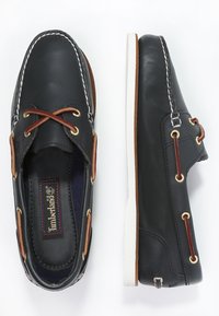 Scarpe da barca da uomo in pelle nera con suola in gomma bianca, lacci marroni, occhielli dorati e cuciture a contrasto lungo i bordi e la linguetta.