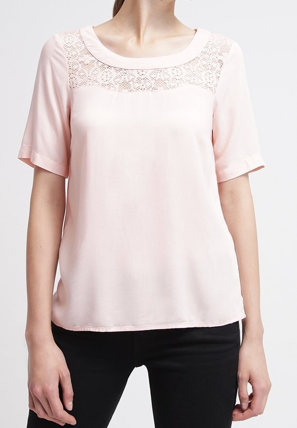 Femme portant un chemisier rose pâle à manches courtes avec un détail en dentelle sur le haut de la poitrine et un pantalon noir.