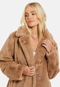 Manteau en fausse fourrure beige avec un col cranté, coupe oversize, fermeture à boutons à l'avant et texture douce. Détails remarquables incluant des accents de boutons brillants.
