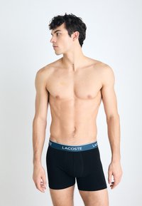 Fekete boxeralsó kék derékkal, amelyen a Lacoste logó látható. Az anyag sima és illeszkedő, a kényelem érdekében tervezték.