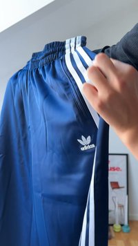 Mão segurando calças de treino Adidas azuis com riscas brancas e logo, em ambiente interior com um poster emoldurado e pesos de ginástica ao fundo.
