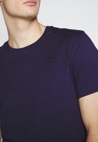 Mies, który nosi gładką ciemnofioletową koszulkę z krótkim rękawem z małym czarnym haftowanym logo na piersi, patrzy lekko na bok.