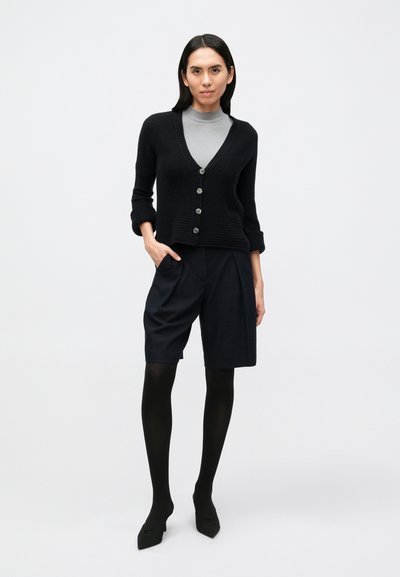 Fekete kötött kardigán V-nyakkivágással és öt gombbal, sötét rövidnadrággal és fekete harisnyával párosítva, hegyes fekete magassarkú cipőkkel kiegészítve.