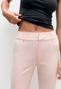 Pantalon rose pâle ajusté avec une texture lisse, taille mi-haute et ceinture traditionnelle. Associé à un haut noir ajusté.