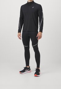 Haut de sport noir à manches longues avec zip moitié, associé à des leggings noirs avec des accents blancs et des chaussures de course noires avec des détails rouges.