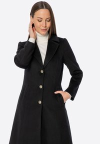 Czarny wełniany płaszcz z wyciętym kołnierzem, pięcioma złotymi guzikami, bocznymi kieszeniami i dopasowanym krojem. Noszony na białym golfie.