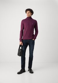Pull en col roulé violet, coupe slim, manches longues, associé à un jean bleu foncé et des chaussures noires. Tient un petit sac bandoulière noir.