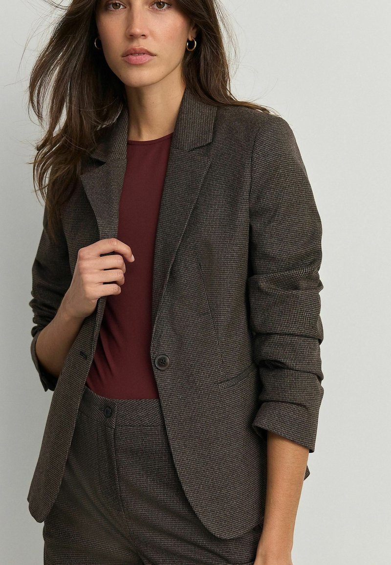Blazer à carreaux marron avec deux boutons, revers en pointe et manches retroussées, associé à un haut bordeaux. Tissu lisse avec un design structuré.
