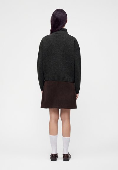 Personne avec de longs cheveux foncés portant un pull gris foncé, une jupe marron, des chaussettes blanches et des chaussures marron foncé, debout de dos sur un fond uni.