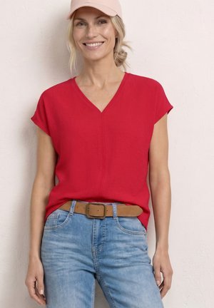 Femme souriante portant une casquette rose, un haut rouge à manches courtes avec un col en V, un jean bleu clair et une ceinture marron, debout contre un mur pâle.