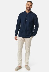 Chemise bleu marine à manches longues avec une patte de boutonnage, associée à un pantalon beige et des chaussures à enfiler de couleur claire, se tenant contre un fond blanc.
